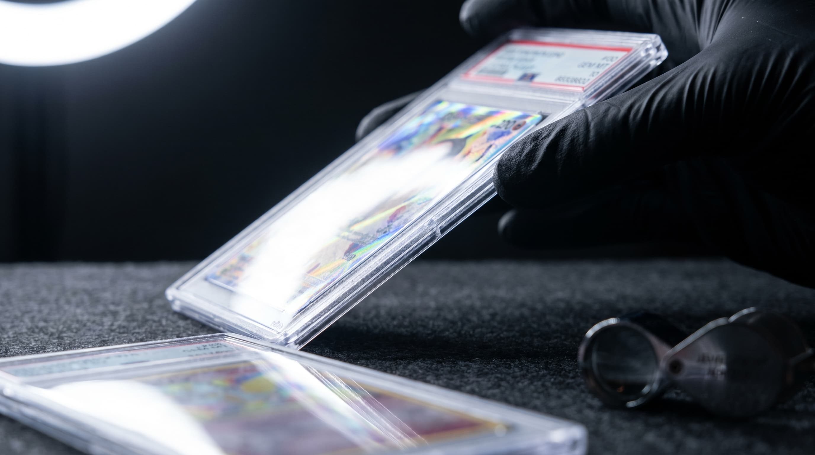 A gloved hand tilts a graded trading card slab so raking light catches a streak of foil colour on the card inside, with a second slab and a loupe blurred on dark felt nearby.