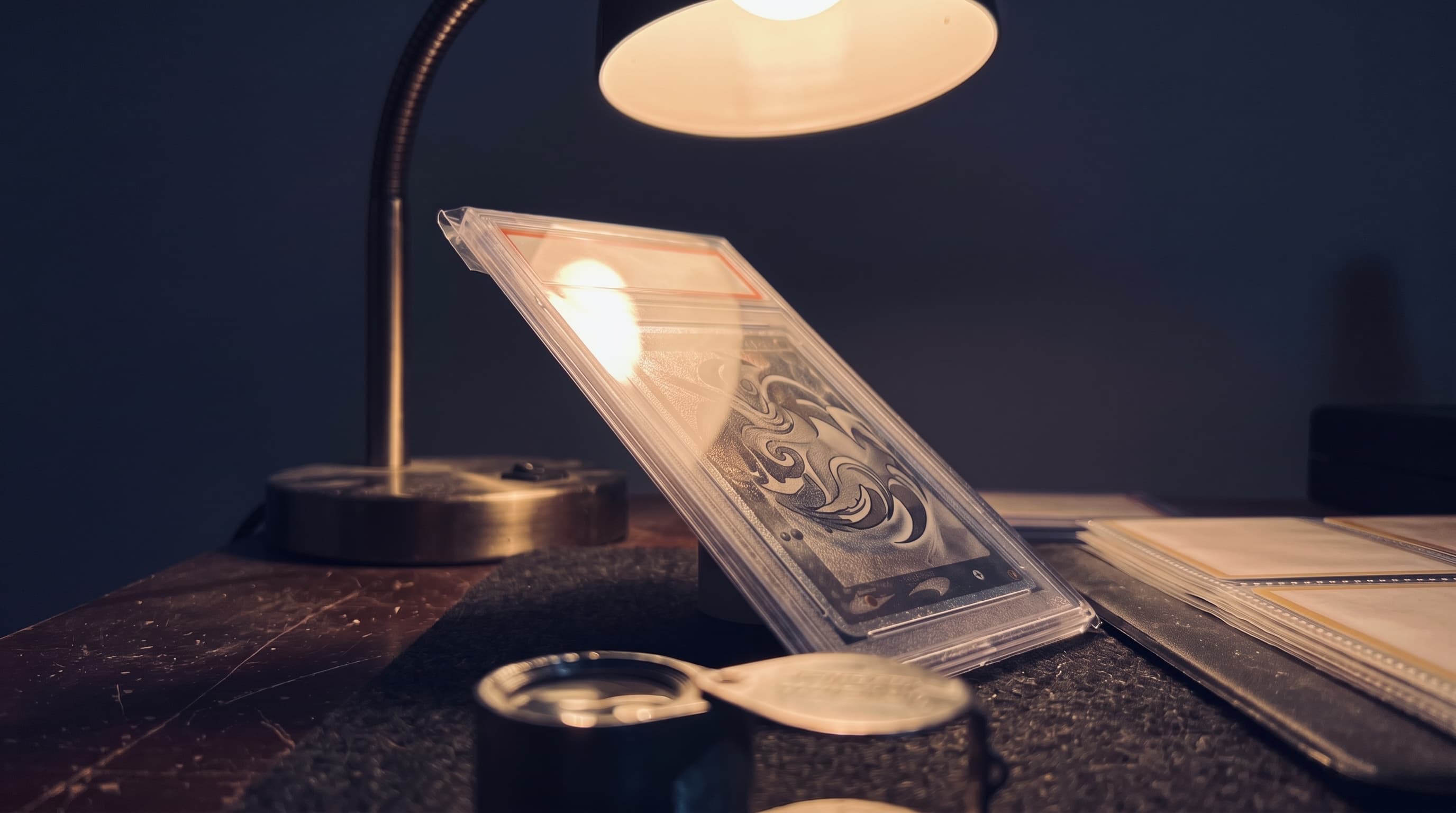 A graded trading-card slab tilted under a desk lamp at night, foil card art catching a streak of warm light, with a loupe and sleeved binder page softly out of focus at the edges.